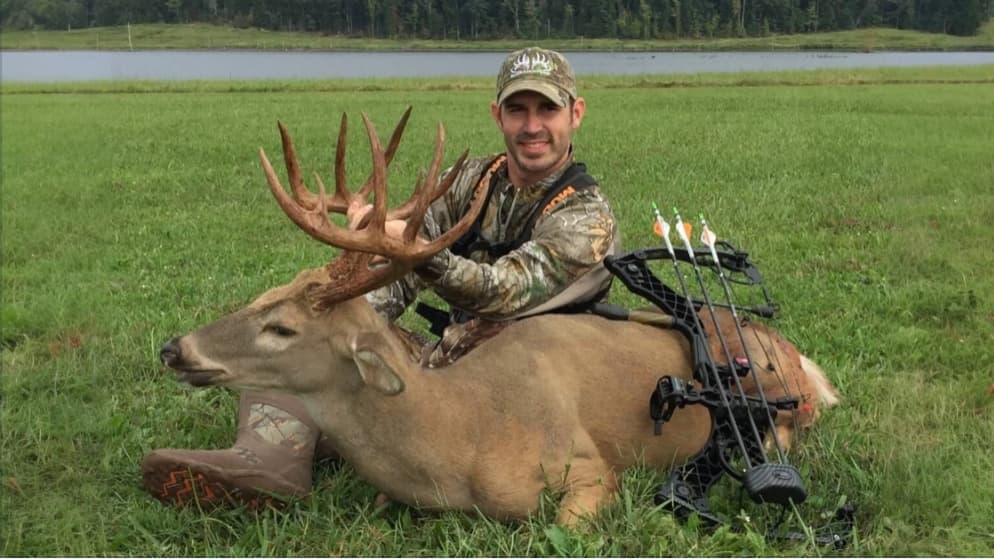 Man in camo sits behind a large buck with antlers on a grassy field. A bow with arrows is visible. Hunter is smiling, out...
