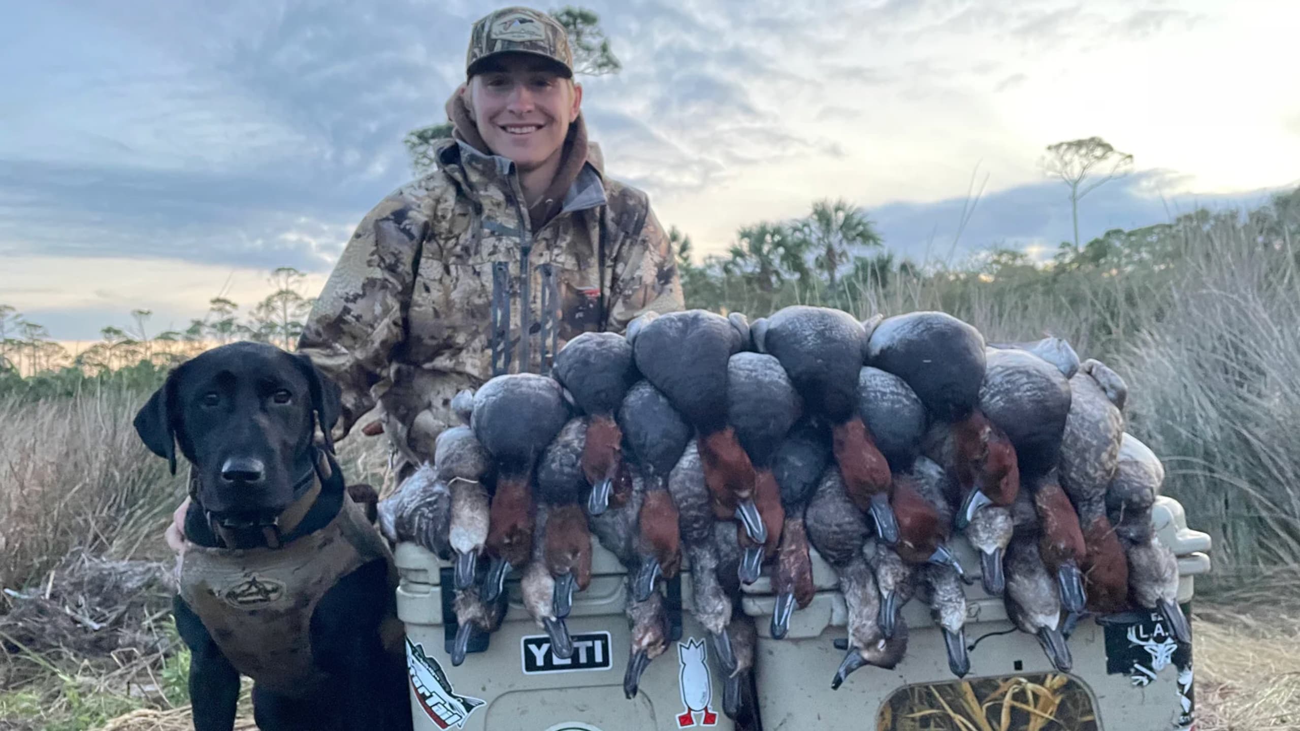 A young man in camo stands next to a black lab. A cooler is overflowing with harvested ducks. Cloudy sky and grassy backgr...