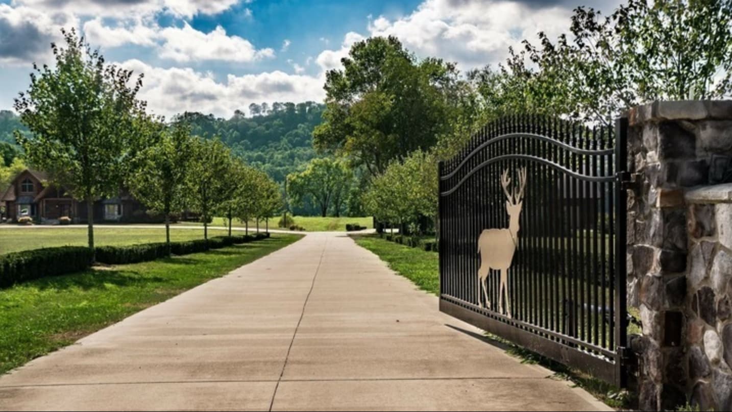 Driveway leading to a house, framed by trees and a decorative gate with a deer silhouette. Green hills and blue sky in bac...