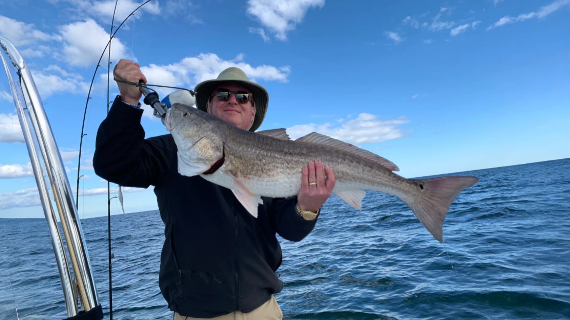 Man on boat holding large redfish. Blue sky and water in the background. Fishing rod visible. He wears sunglasses and hat.