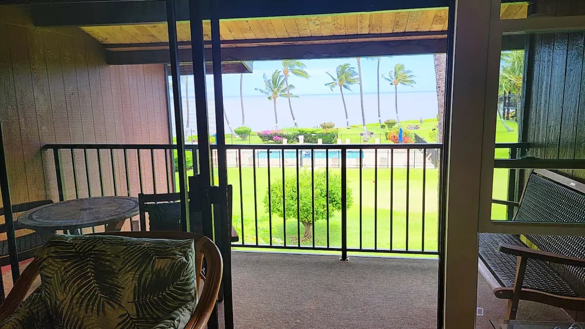 View from balcony overlooking palm trees, ocean, and a lawn. Furnishings on the balcony include a table, chairs, and bench.