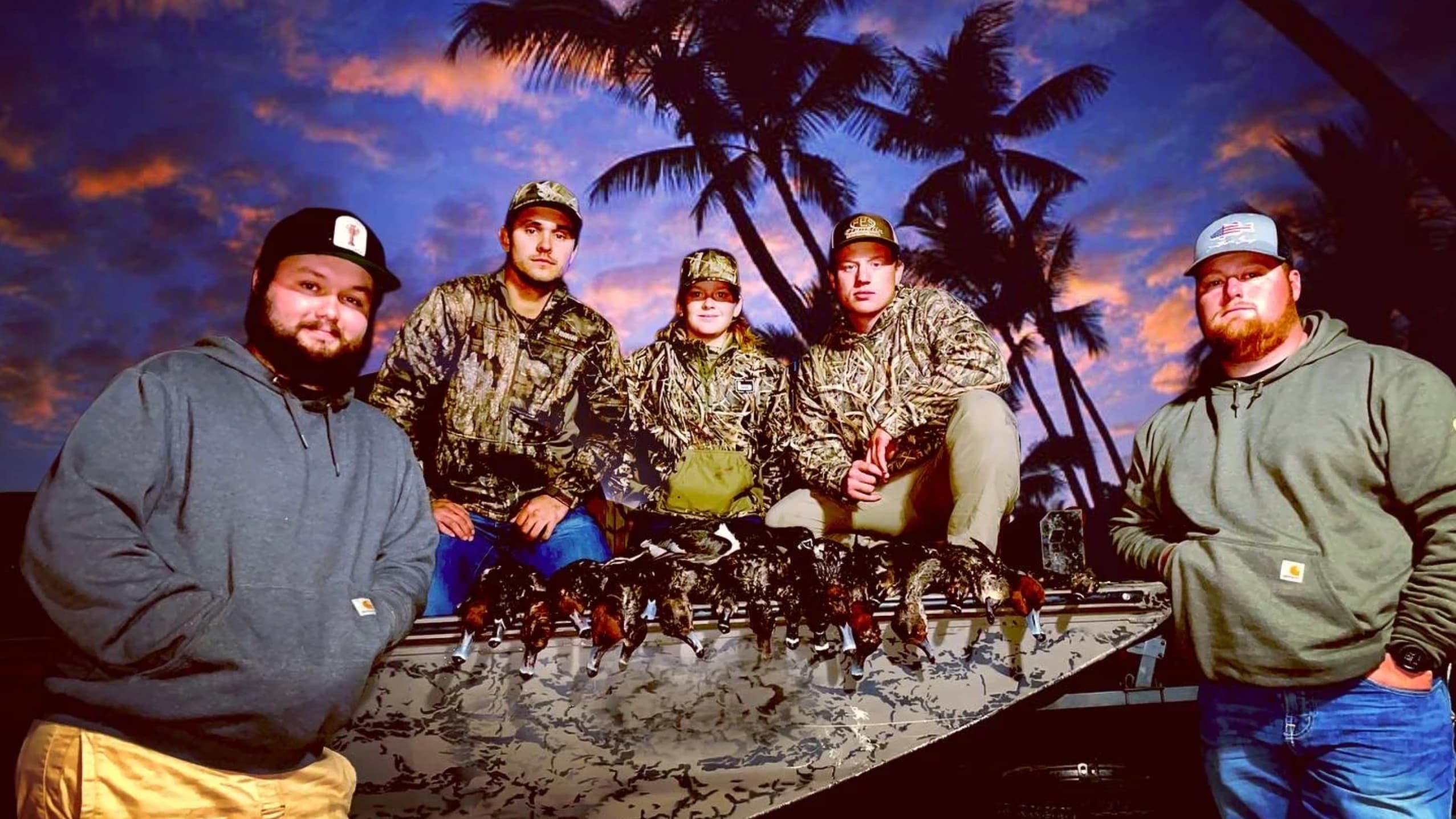 Five people pose with harvested ducks on a camouflaged boat. All wear hats and camouflage jackets. Ducks hang from the boa...