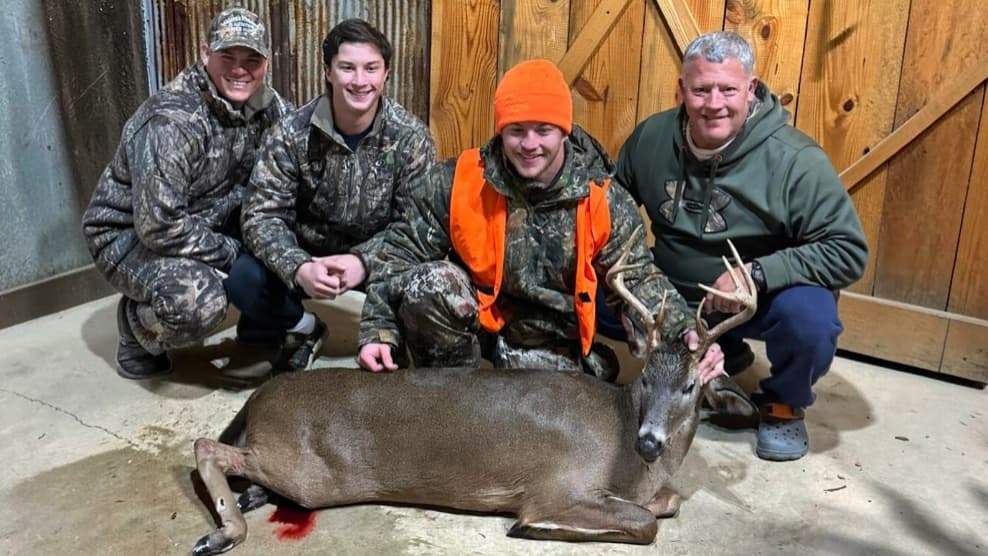 Four men kneel with a dead deer. The man in the middle wears an orange vest and hat. The others wear camo or green, with t...