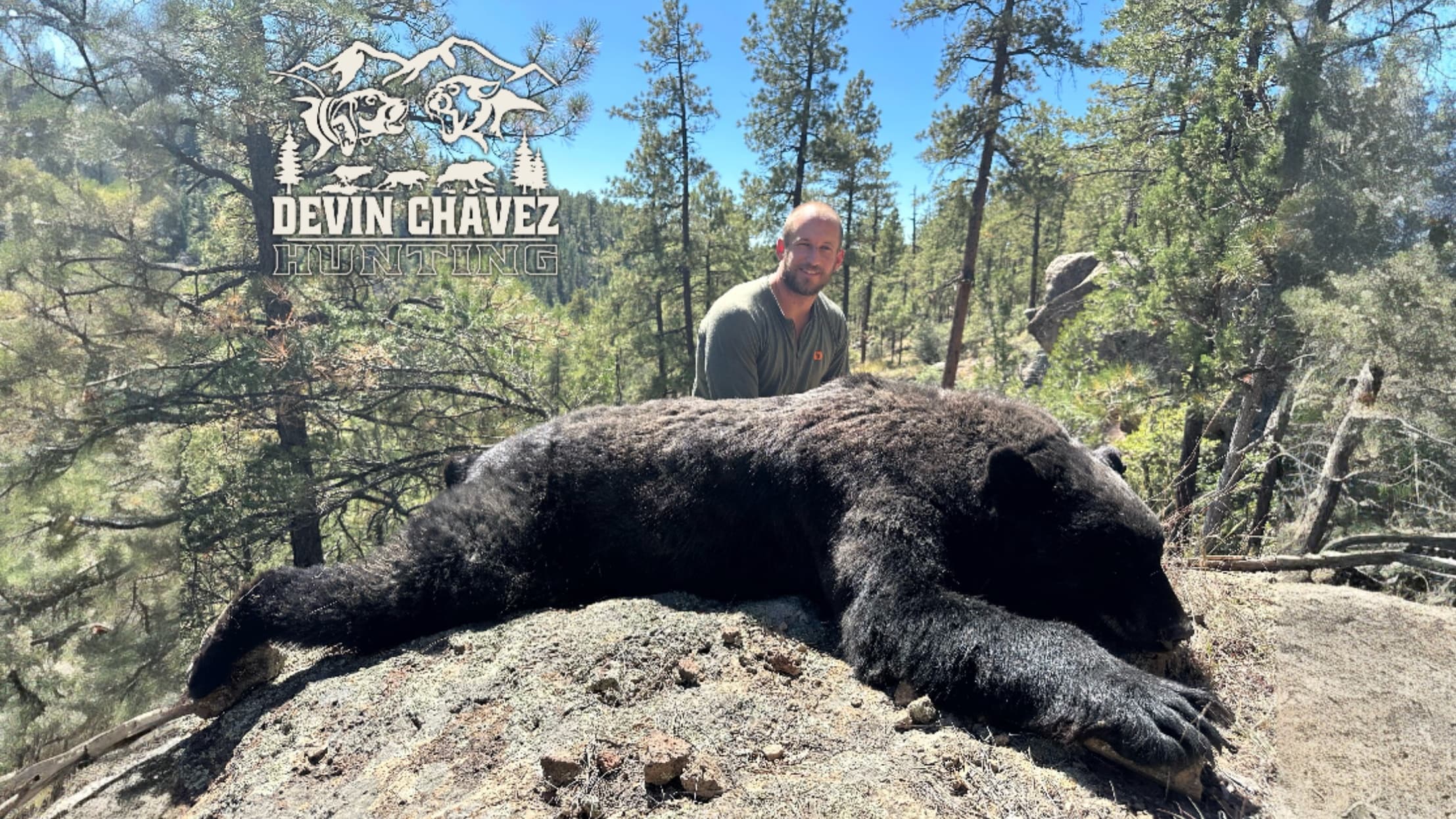 A man stands behind a large, black bear lying on a rocky surface. Trees fill the background; hunting logo visible.