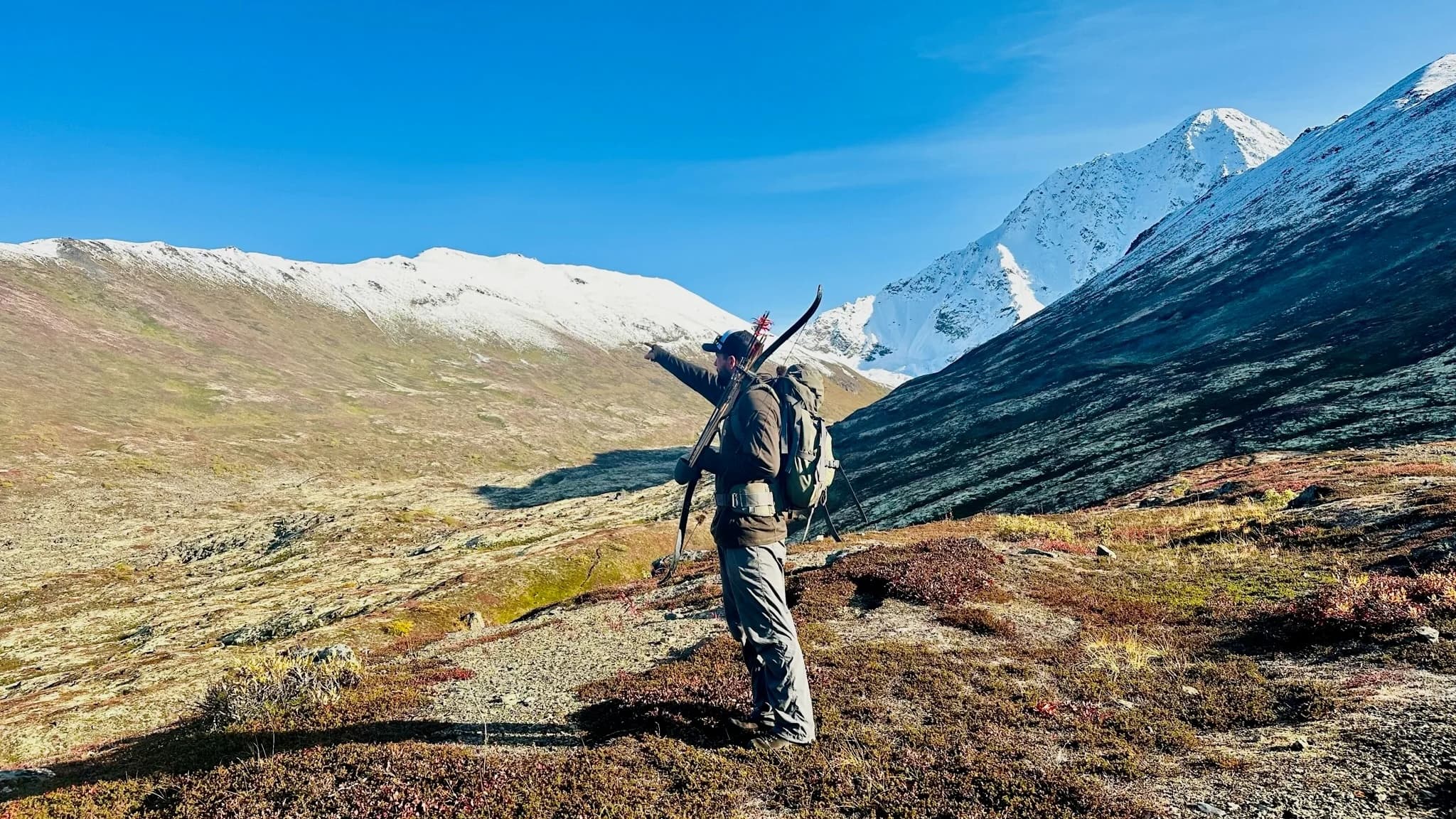 Person with bow and backpack points towards snow-capped mountains. They stand on a hill with sparse vegetation under a br...