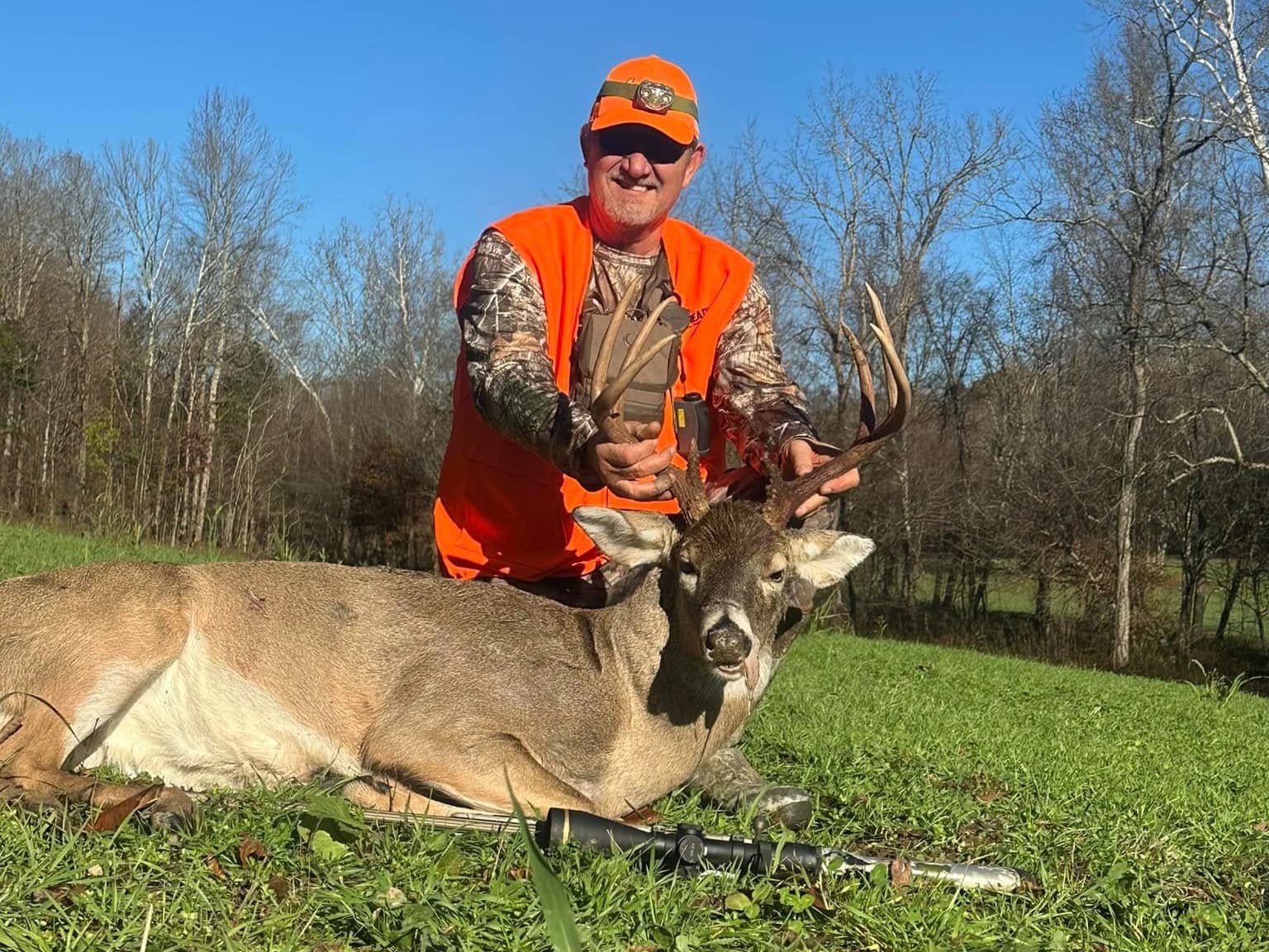 Hunter in orange vest holding large buck with antlers. Deer lies on green grass, rifle beside it. Background is trees and ...