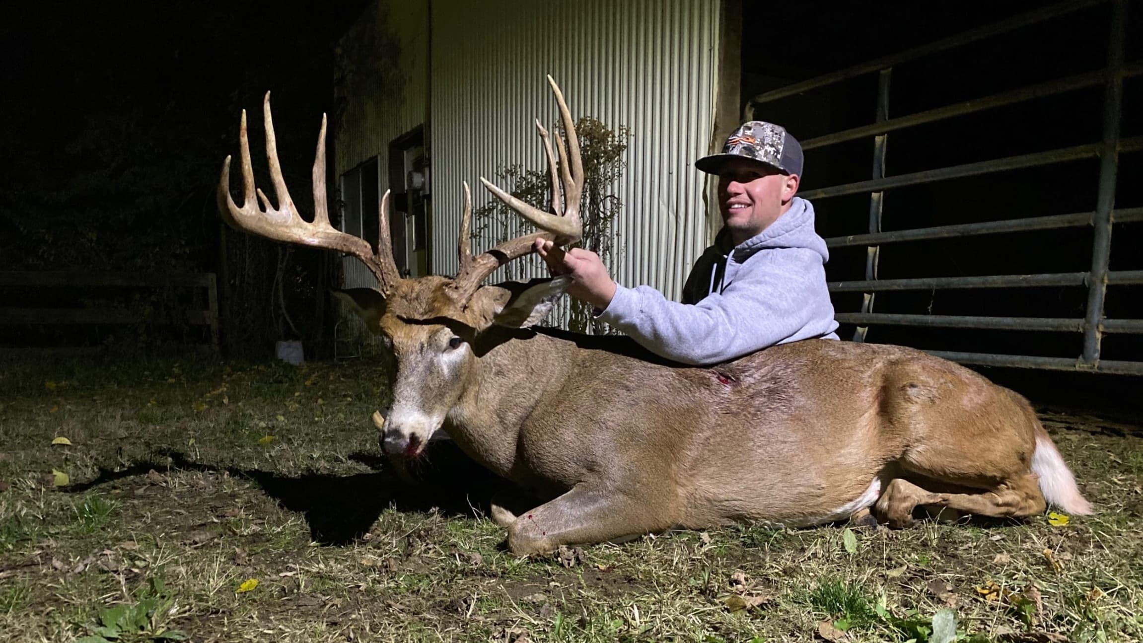 Man posing with a large buck. The deer lies on the ground with large antlers, a man is smiling and holding the antlers. N...