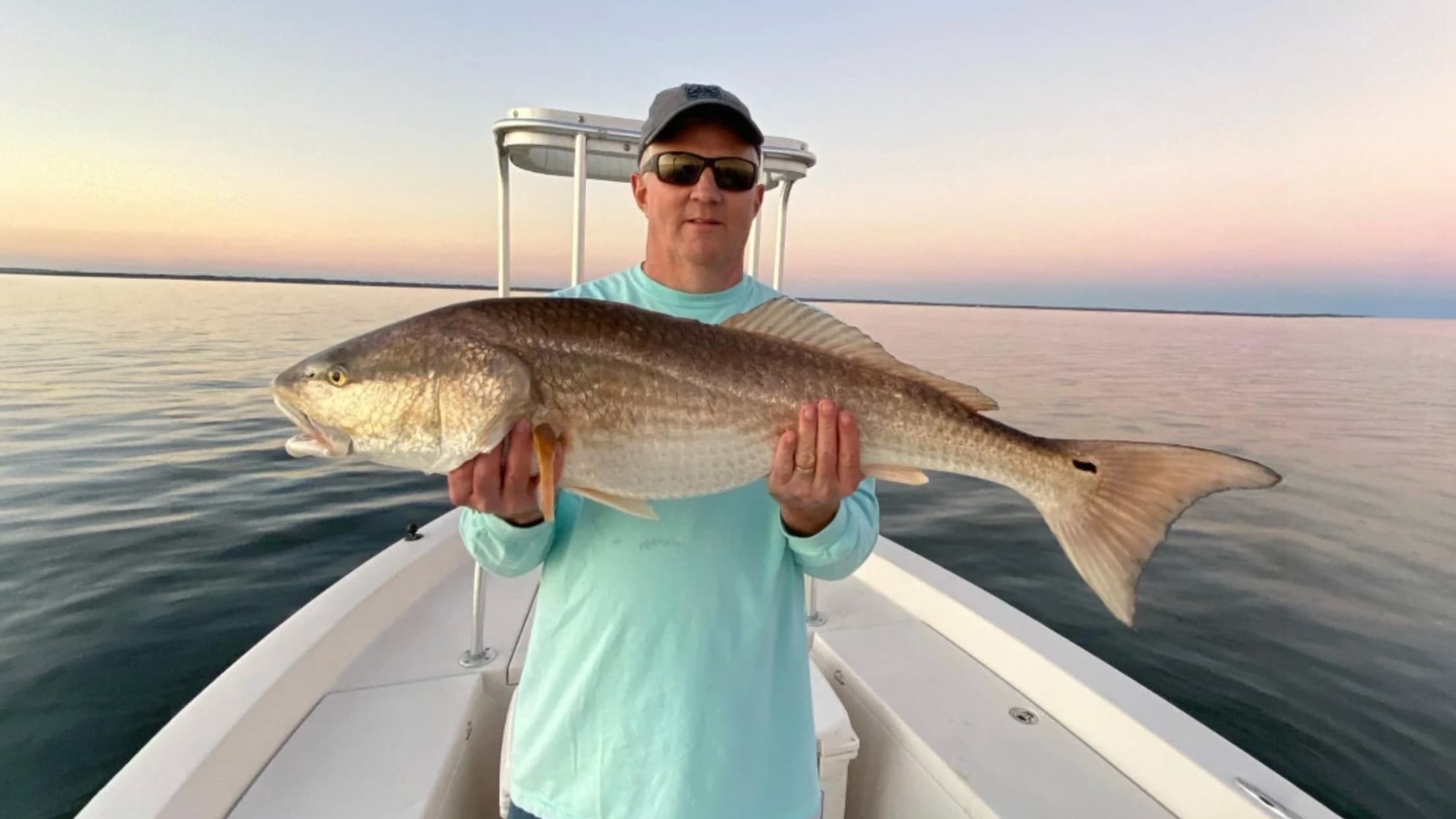 Man on boat holding large redfish. Sunset sky, calm water. Man wearing hat, sunglasses, light blue shirt. Fish is reddish-...