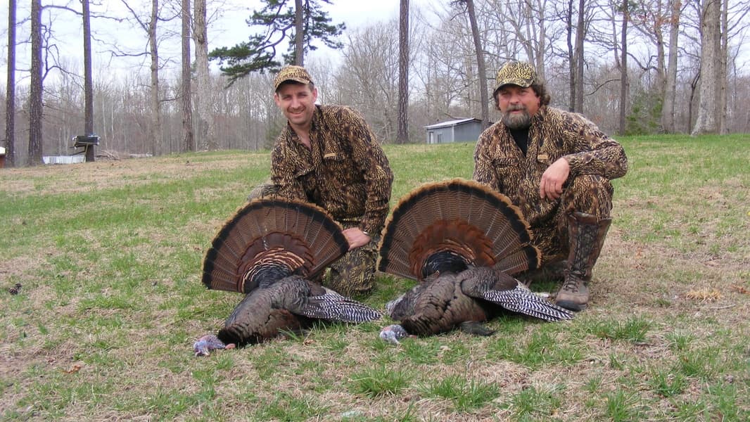 Two hunters in camouflage pose with two wild turkeys, fanned tails displayed, in a grassy, wooded area.