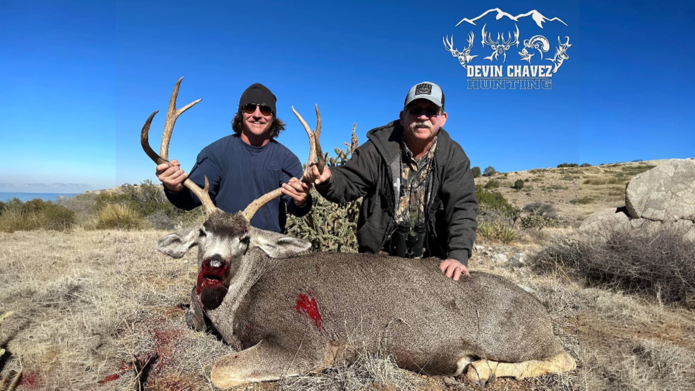 Two men with a large buck, antlers held up. Buck lies on the ground, bloodied. Blue sky, logo says Devin Chavez Hunting.