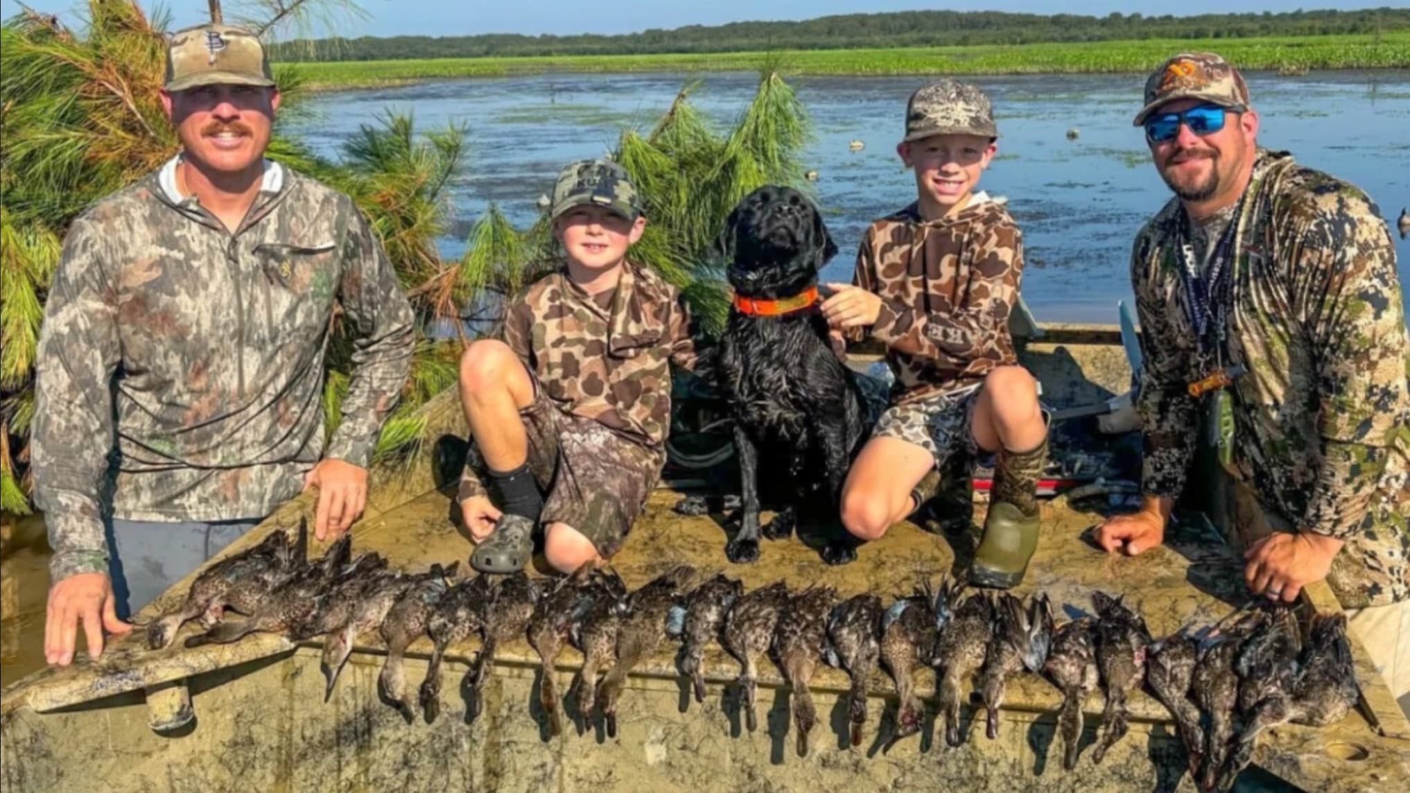 Four people, two boys and two men, pose with a black dog and ducks. They are in camouflage clothing near a body of water. ...