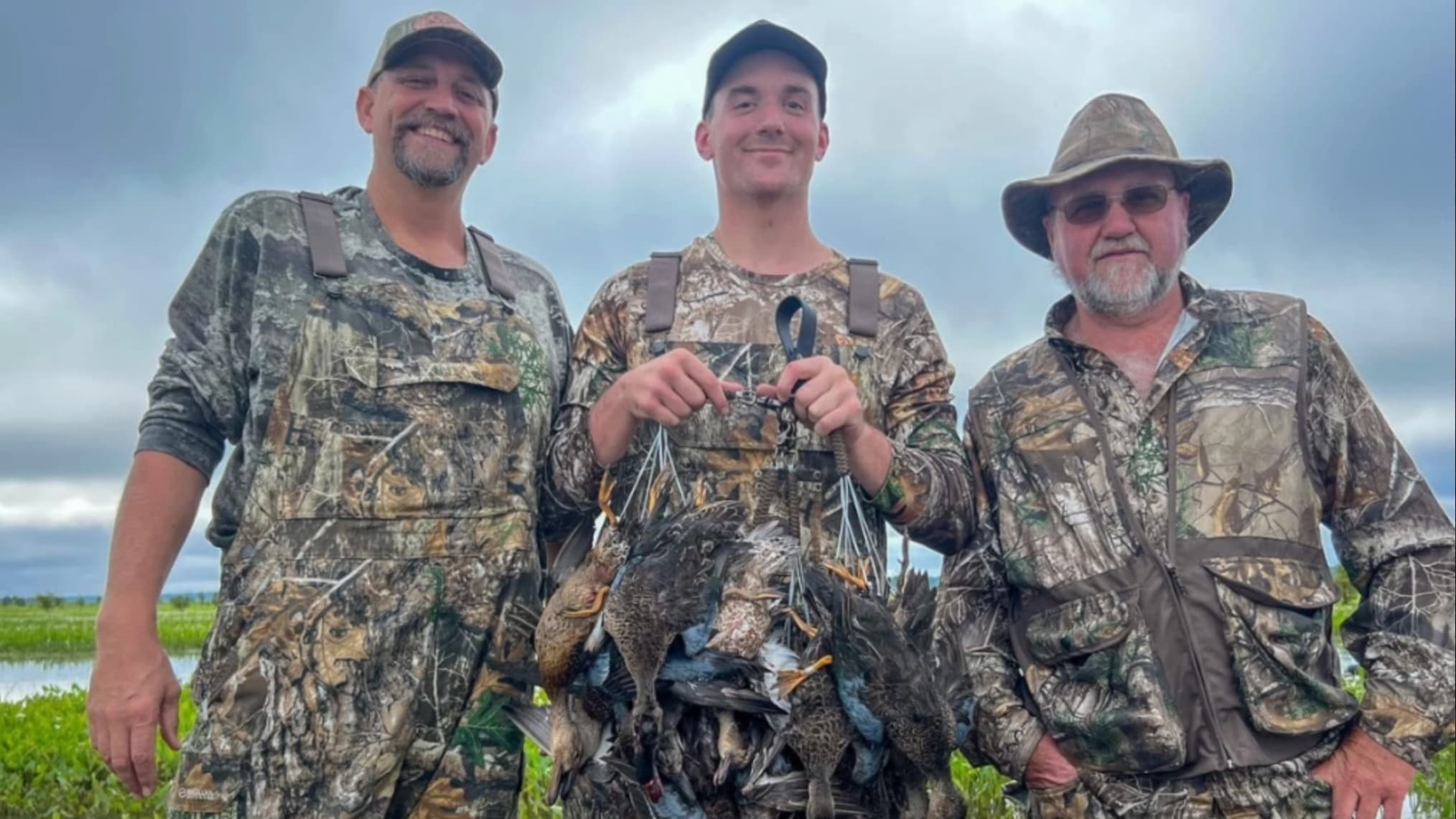 Three men in camouflage gear pose, the central figure holds a string of dead ducks. Background shows marsh, cloudy sky.