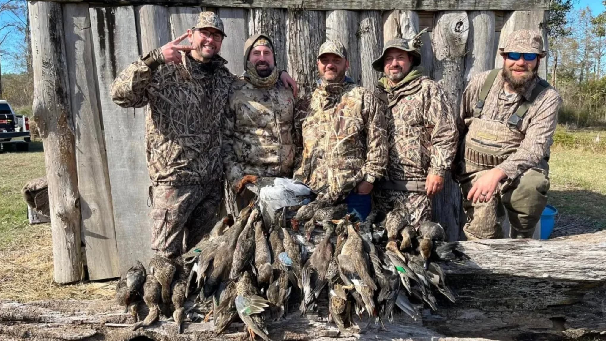 Five men in camouflage pose next to a pile of ducks they hunted. They stand against a wooden shelter. One man makes a pea...
