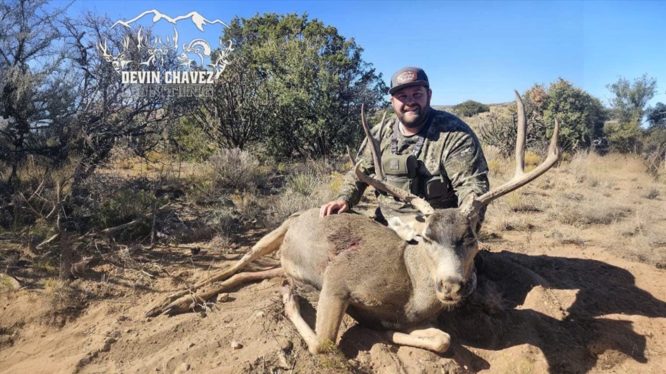 Man with camo clothing kneels beside a large buck with antlers on the ground. A wound is visible on the buck. Blue sky, dr...