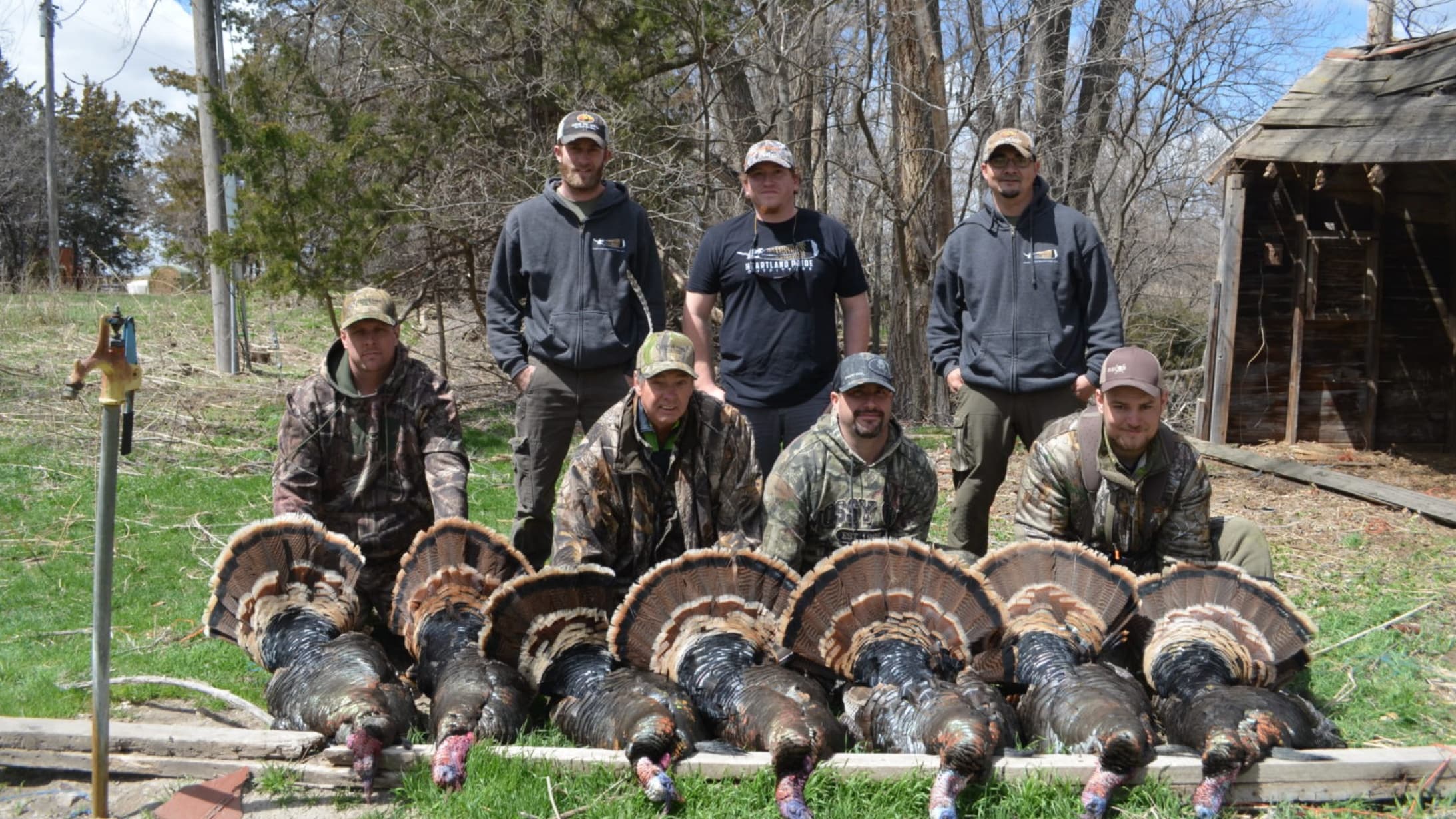 A group of eight men with harvested turkeys. The turkeys are displayed in a row on a wooden platform. The men wear camo an...