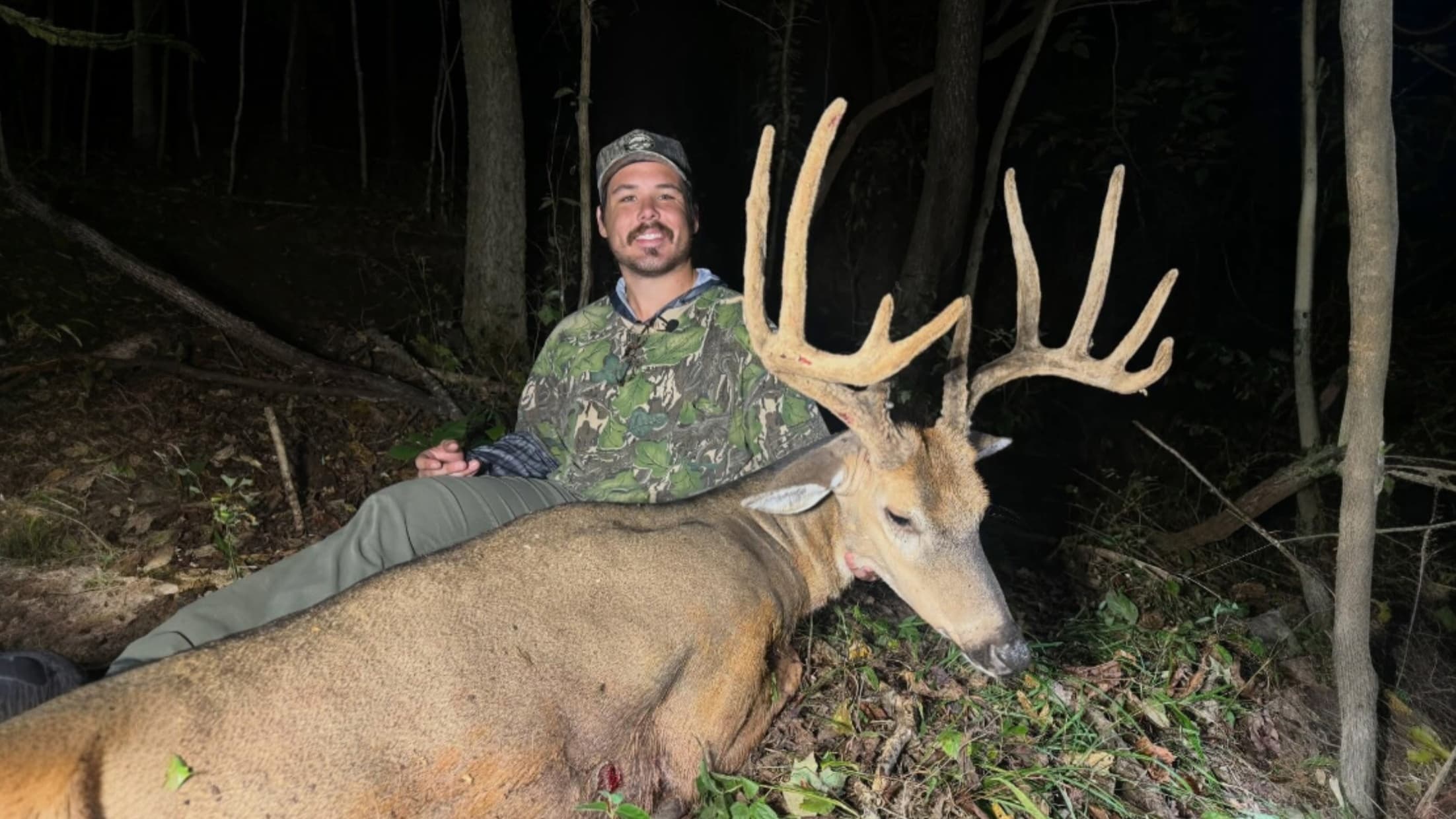 Man in camo next to a large buck with impressive antlers. Deer lies on the forest floor, shot in low light. Man smiles at ...