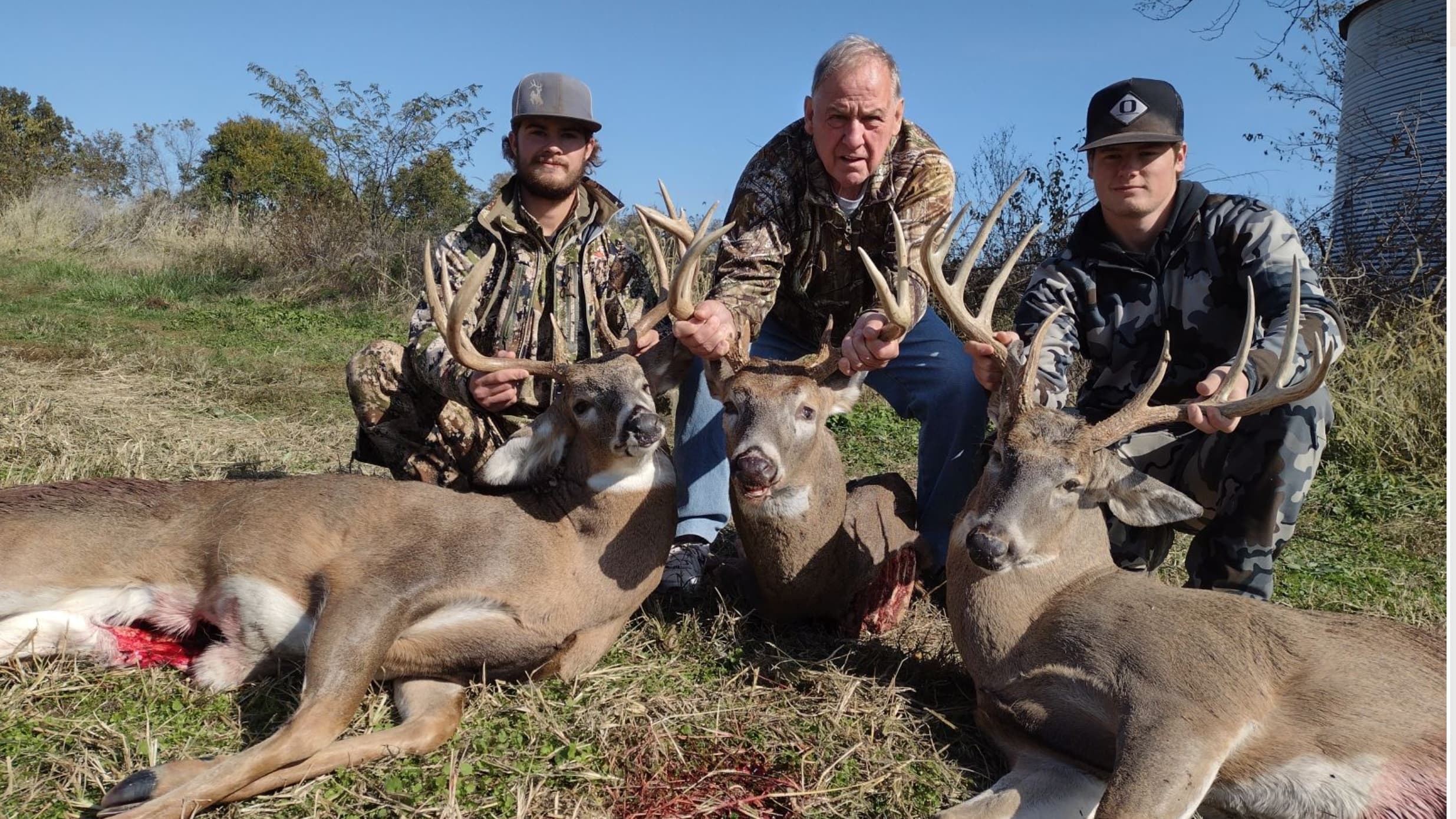 Three hunters show off three large deer carcasses outdoors under a clear blue sky.