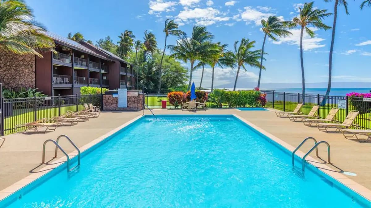 A rectangular pool with clear blue water. Palm trees, a resort building, lounge chairs, and ocean views surround it. Brigh...