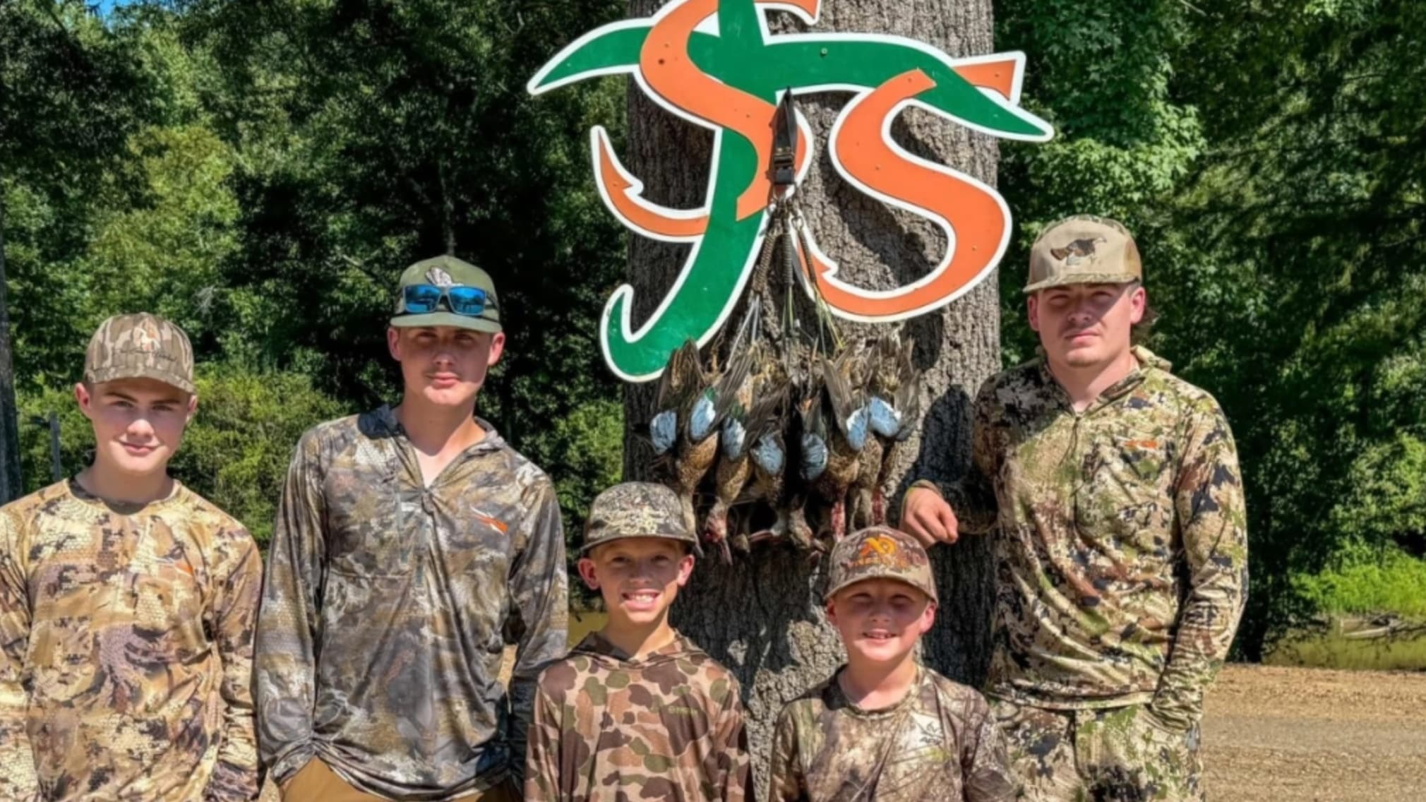 Five young people in camo gear pose in front of a tree. Several ducks hang from the tree. Sign with green and orange lett...