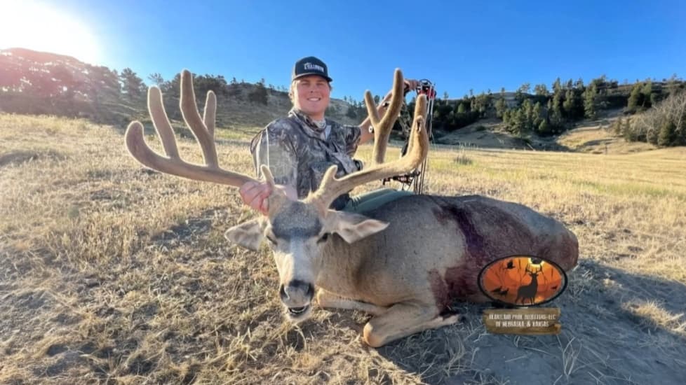 A smiling man in camo holds a large buck's antlers. The deer lays on the ground with a bloody chest. A hunting logo is in ...