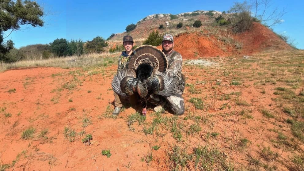 A man and boy pose with a large turkey, its tail feathers fanned out. They kneel on reddish-brown ground, with a hill in b...