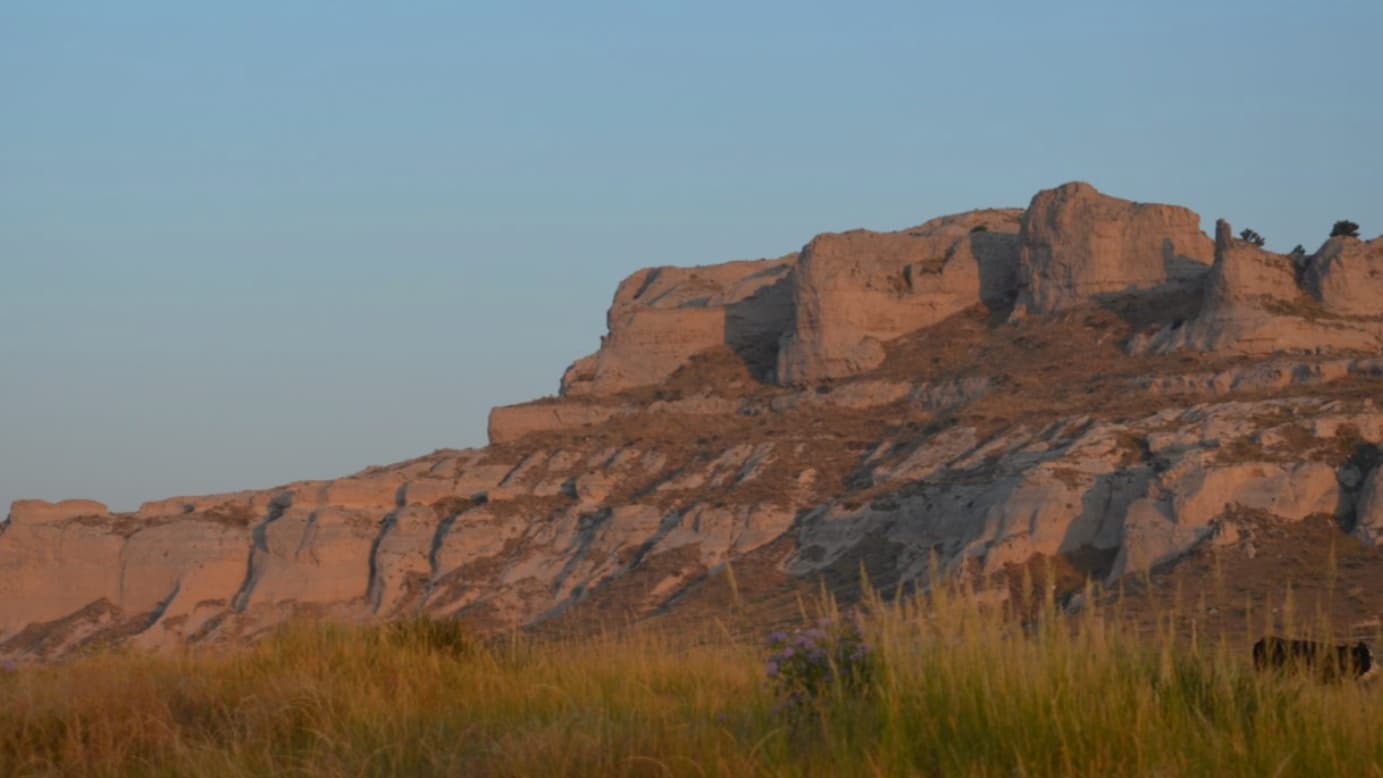 Rocky mesa at dusk, lit with warm tones. Tall grass in foreground, pale blue sky above.