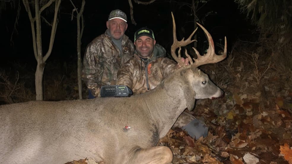 Two men pose with a large buck. The deer lies on leaves with large antlers. Men wear camo and hats, one holds the buck's a...