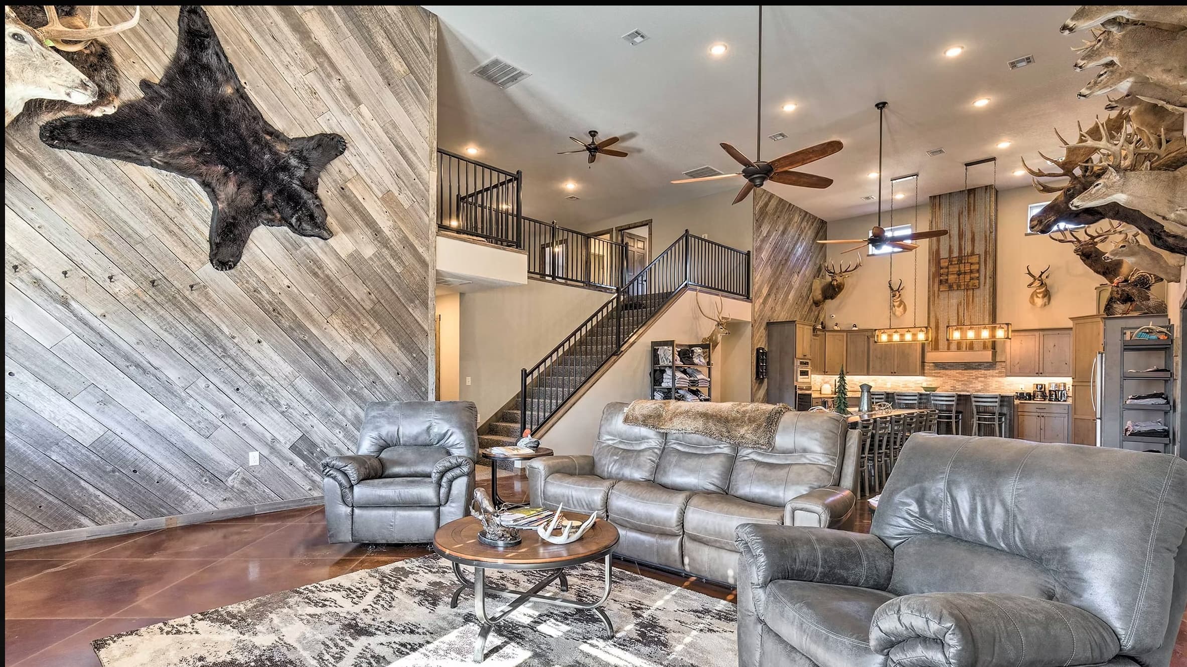 Spacious living room with rustic wood paneling. Bear rug on the wall, leather furniture, and deer antler decor. Kitchen an...