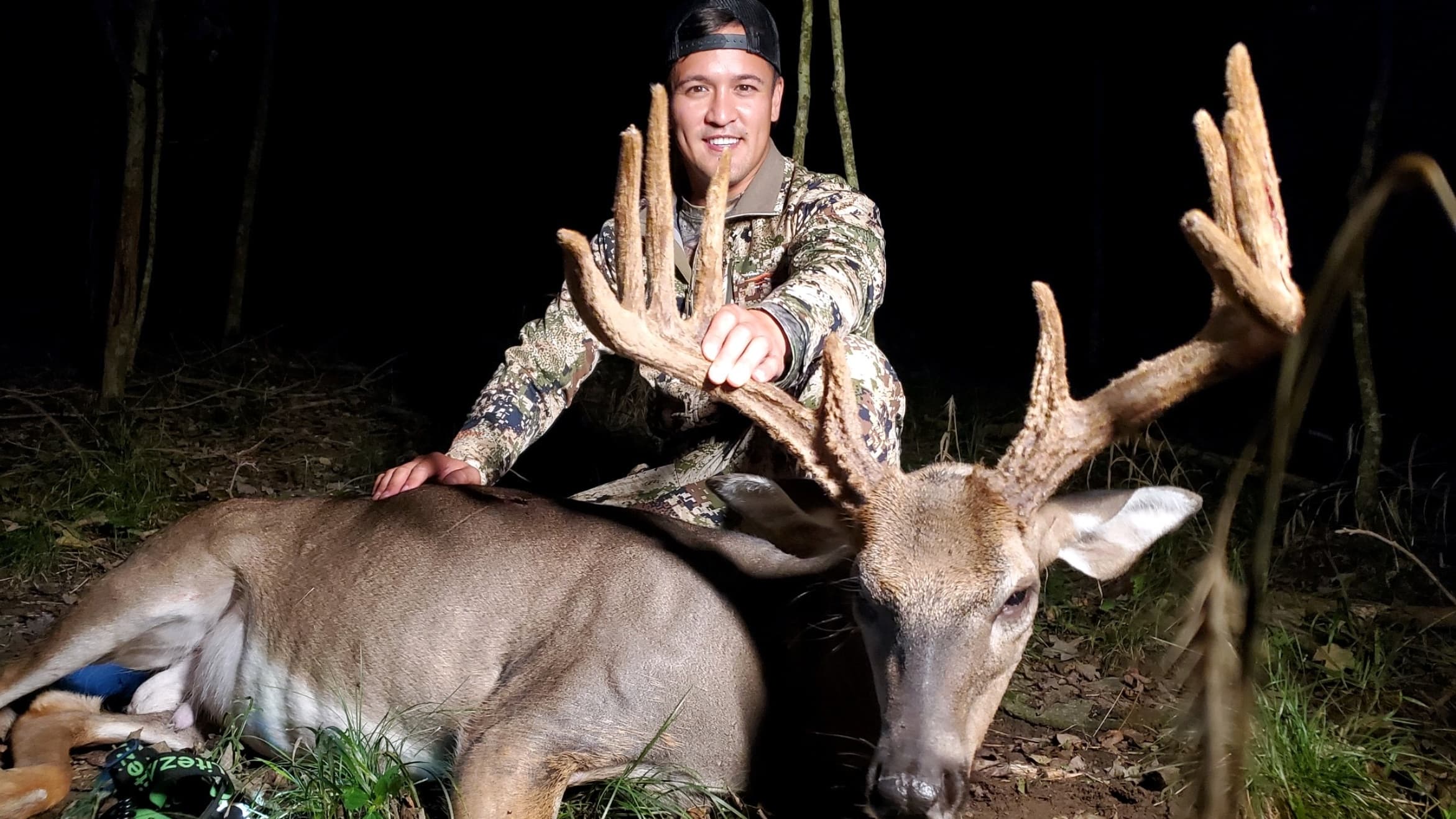 Man in camouflage holds up antlers of a large buck. Buck lies on the ground with the man in a dimly lit forest, night time.