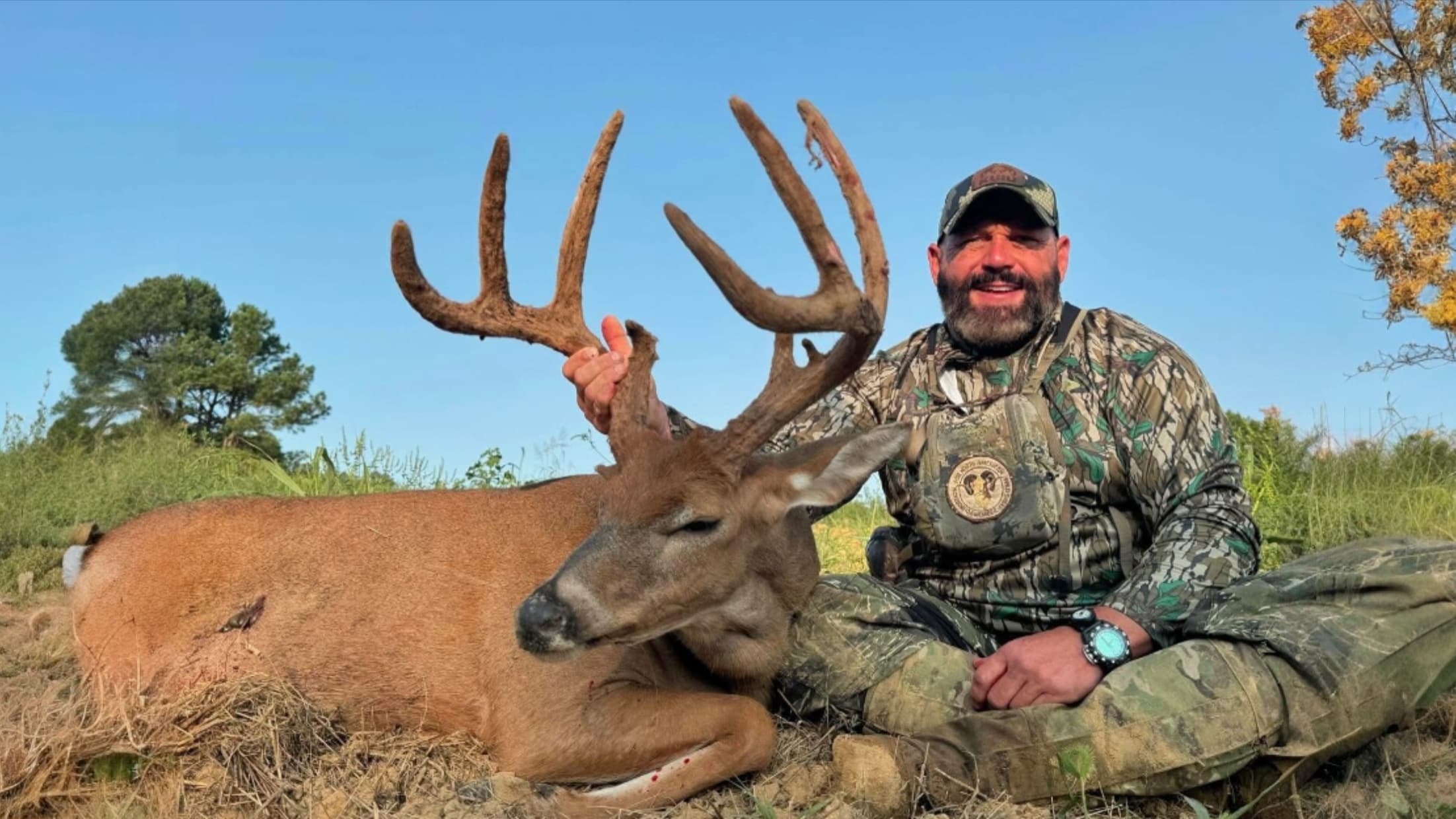 Man in camouflage sits next to a large buck with impressive antlers. Deer rests on the ground, man holds antlers, smiling....