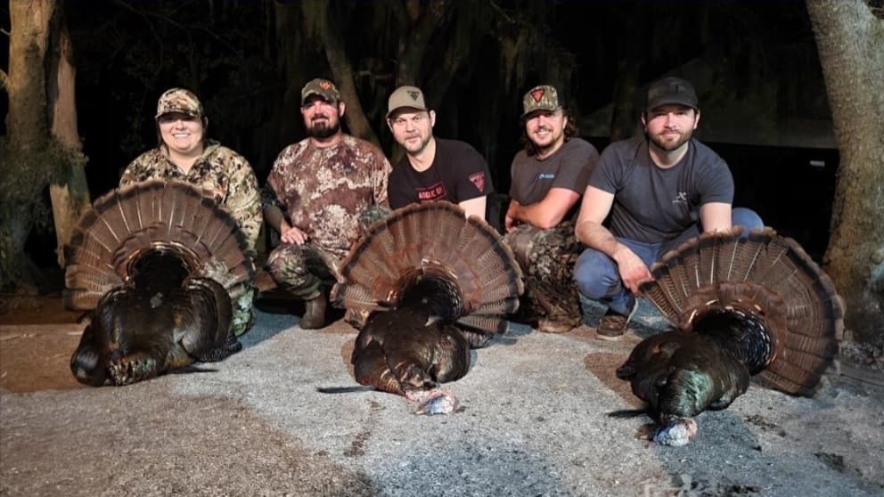 Five people pose with harvested turkeys. Each person kneels behind a turkey displaying its fanned-out tail feathers. Dark,...