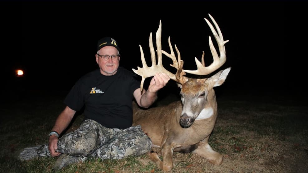 Man with glasses and a black shirt holding large deer antlers, next to a deer lying down. Both are on grass. Dark background.
