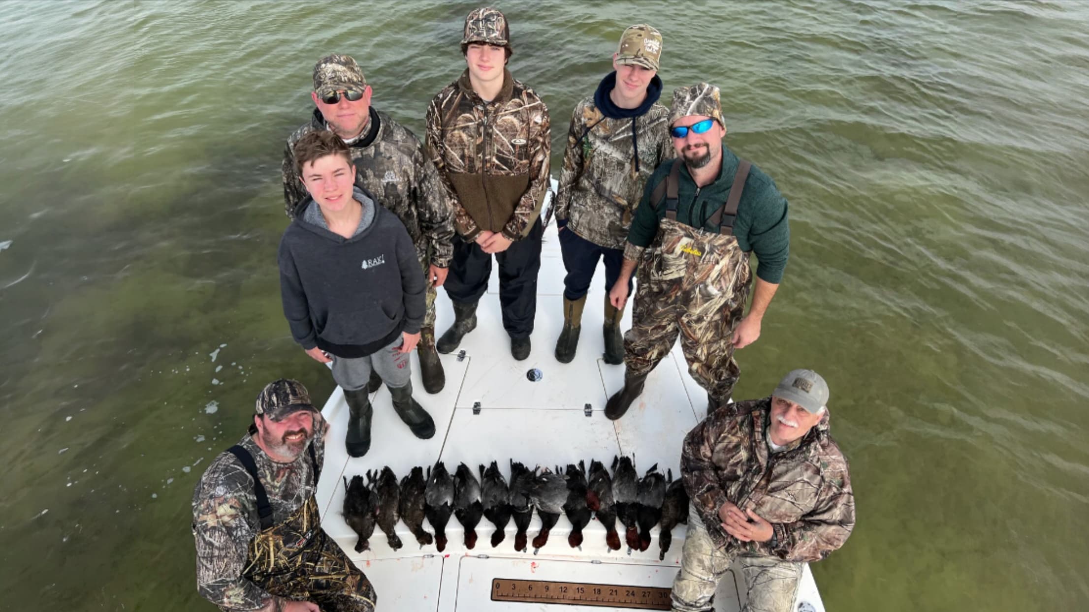 Group of seven men posing on a boat with a row of harvested ducks. Most wear camo gear. Boat floats in calm, green water.