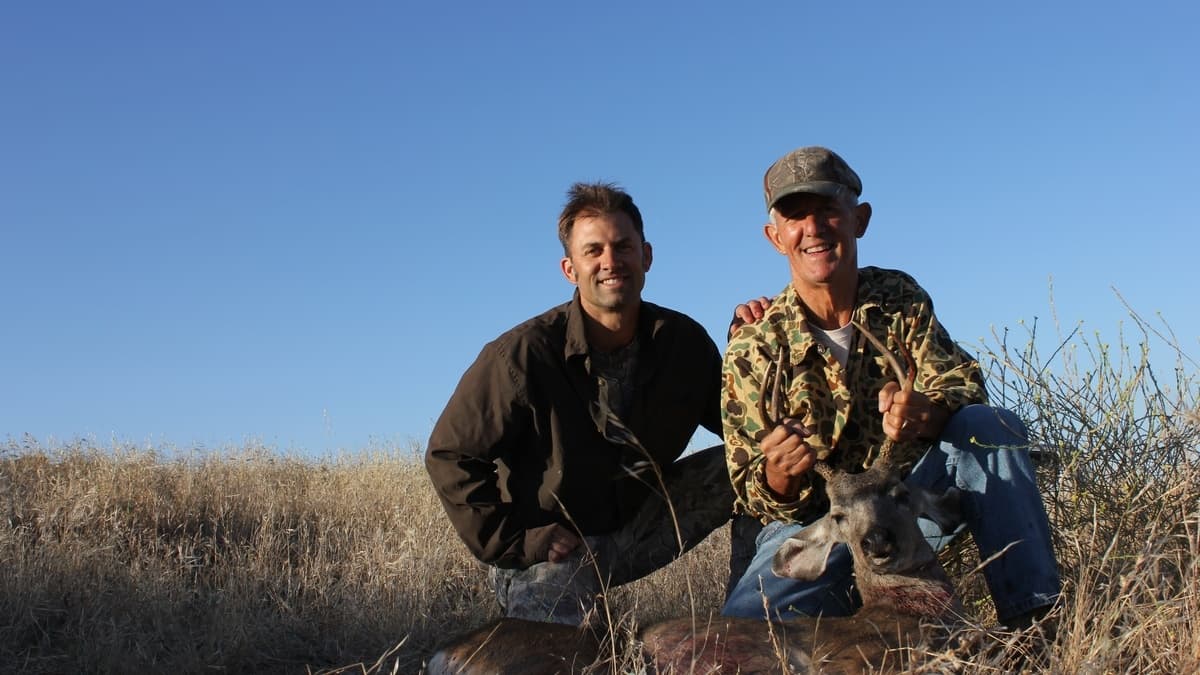 Two men kneel beside a deer they have killed. One holds the antlers, both smile with pride. Blue sky in background.