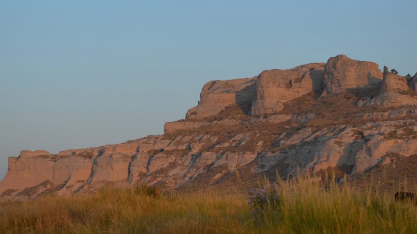 A large, layered rock formation illuminated by golden sunlight. Tall, dry grass in the foreground and a blue sky above th...