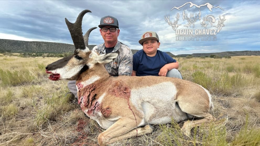 Man and child pose with a harvested pronghorn. The animal lies in dry grass, showing visible wounds. Cloudy sky in the bac...