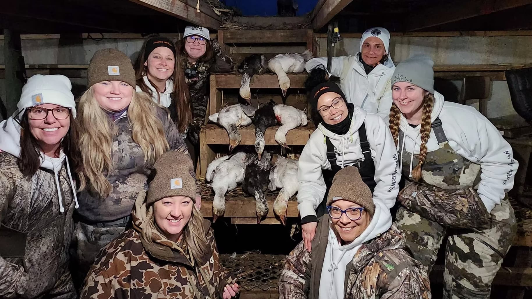 Group of women wearing hunting attire, posing with harvested geese. Geese on wooden shelves, all smiling and facing camera.