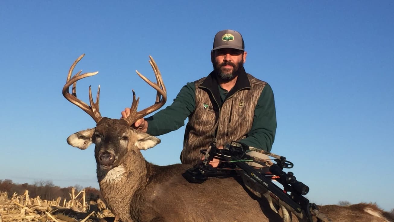 Hunter with large antlers holding a crossbow next to a harvested deer.