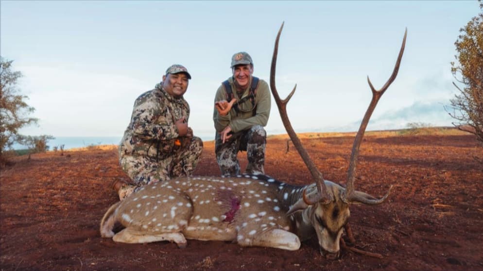 Two men pose with a spotted deer with large antlers on a reddish ground. One man kneels, the other crouches beside it.