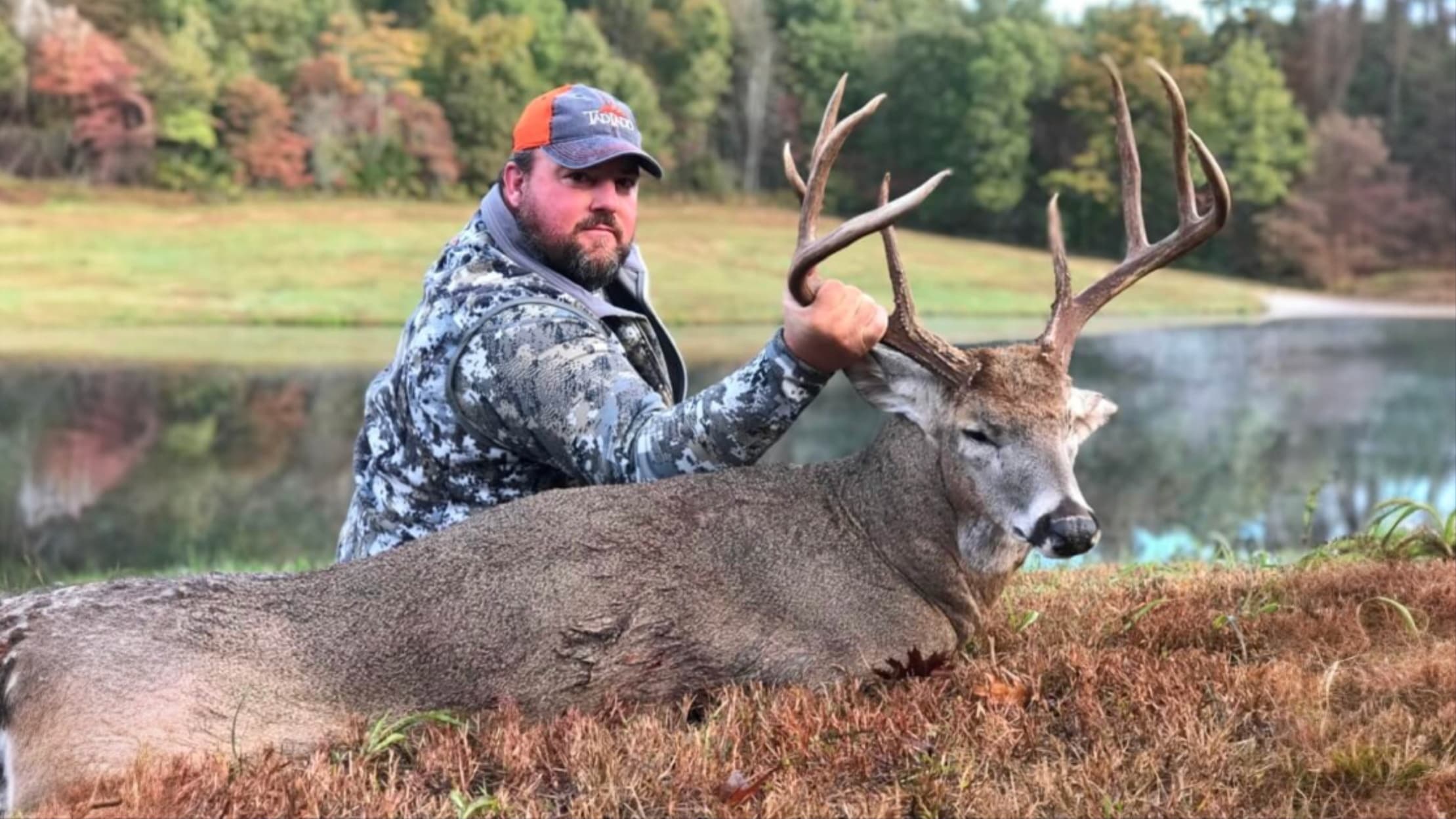 Man in camo jacket and orange hat posing with a large buck deer, antlers visible. Deer rests on dry grass with a pond in t...