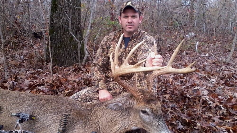 Man in camo kneeling beside a large buck with antlers. He holds the antler, smiling at the camera. Hunting scene in the wo...