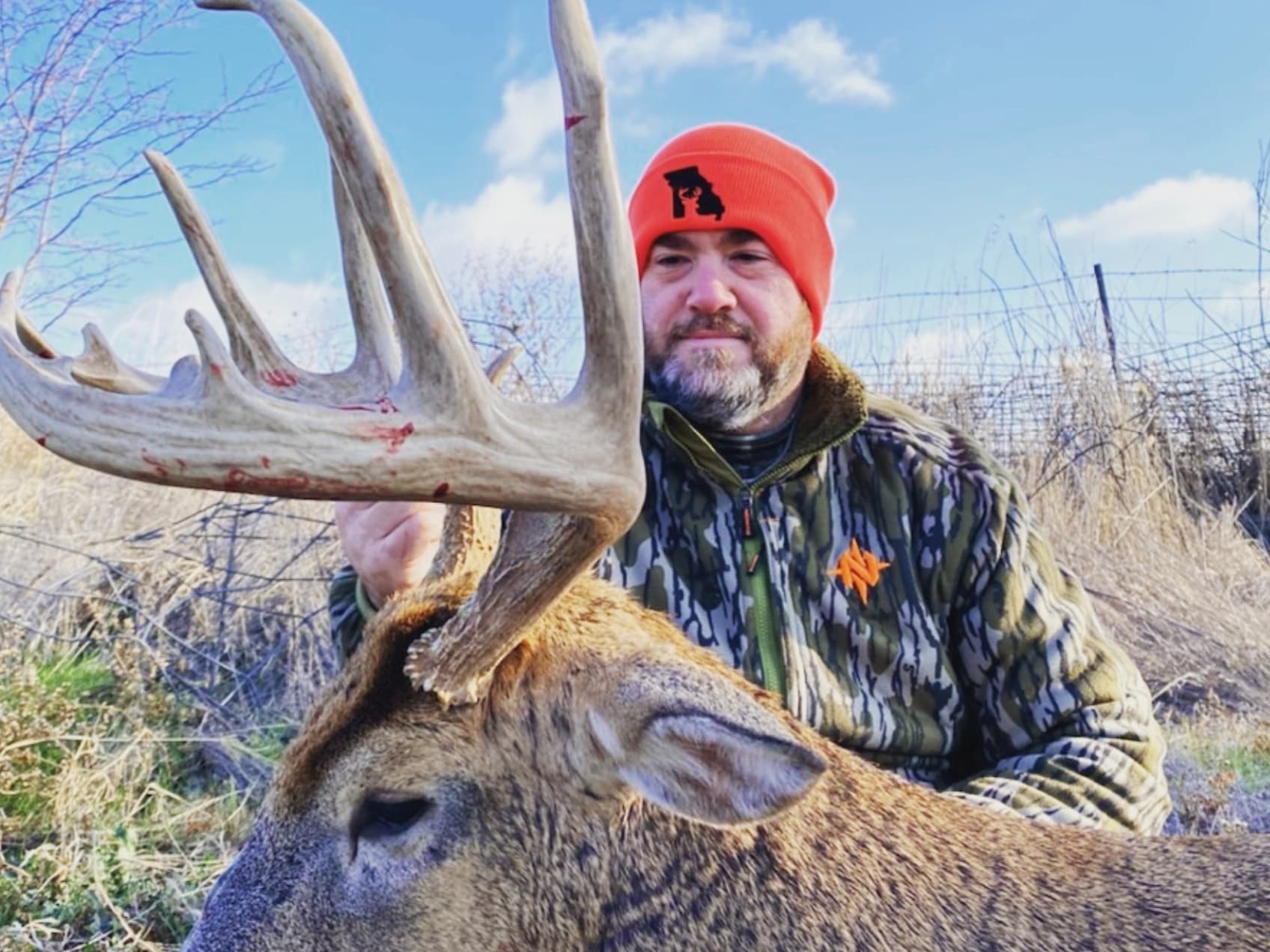 Hunter in orange beanie and camo jacket with a large deer rack with blood.
