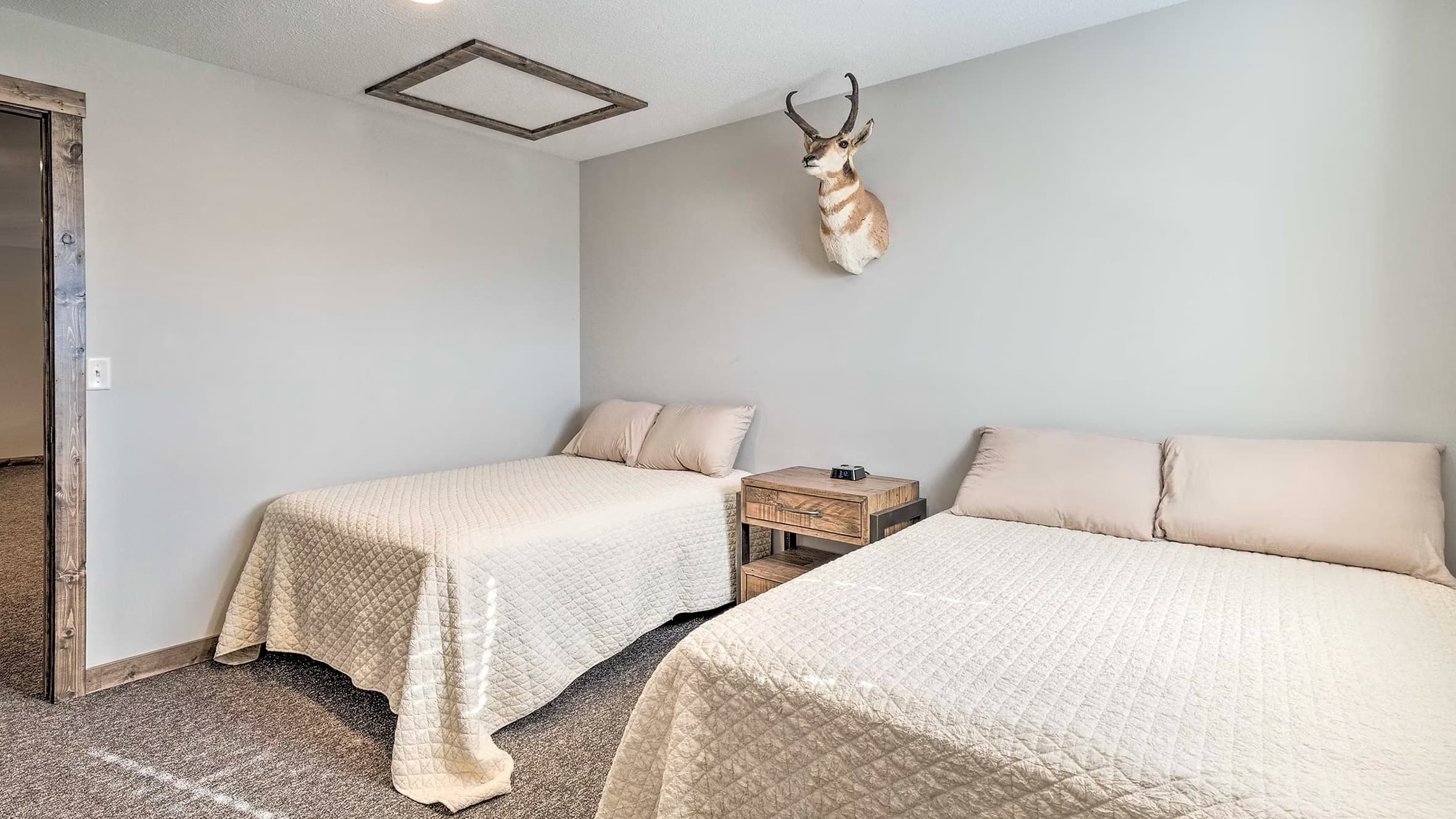 Bedroom with two twin beds, tan comforters, a wooden nightstand, and an antelope mount on wall. Brown trim and carpet.