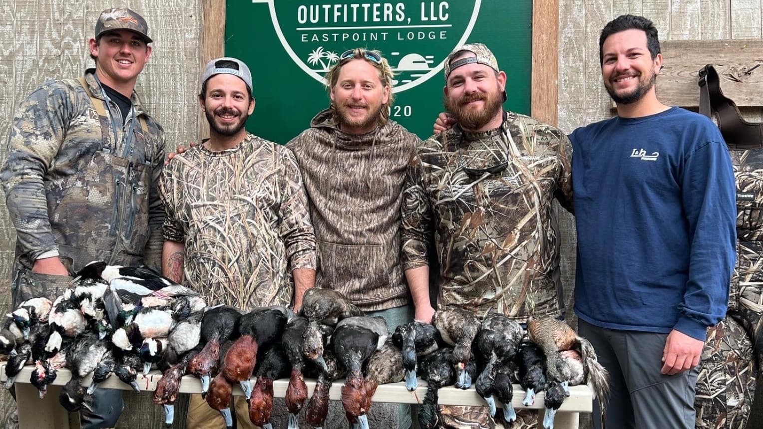 Group of men with harvested ducks. Men in camouflage pose with the birds. Outfitters sign in the background.