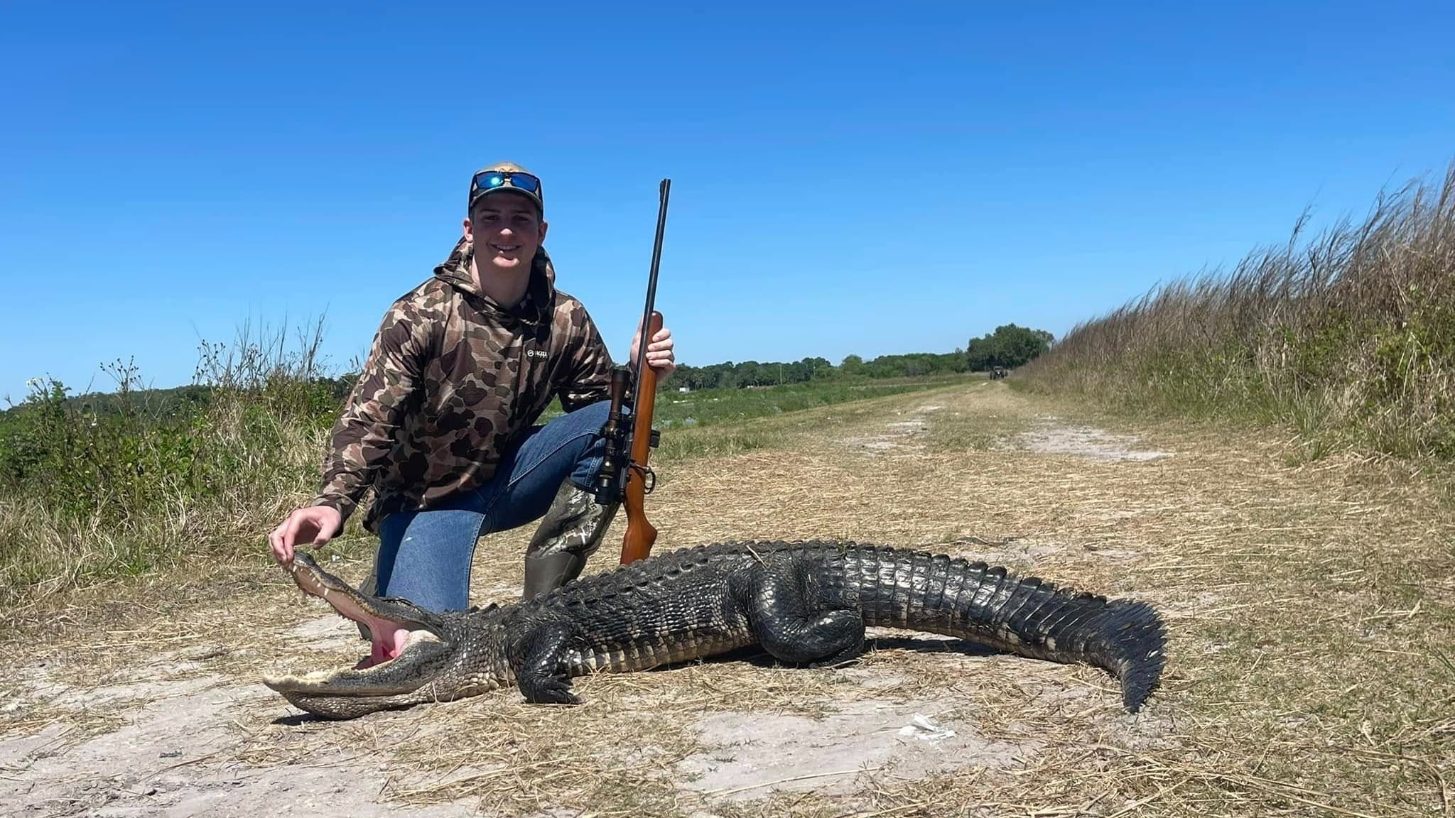 Young man in camouflage holds rifle next to a large alligator with open mouth on a dirt path.