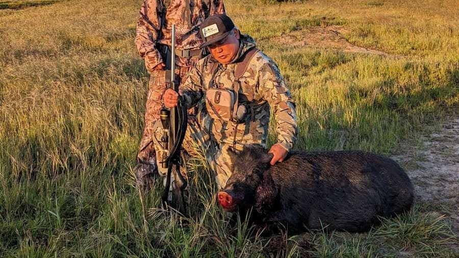 Two hunters with camouflage clothing. One touches a large, dead black hog with blood on its snout. One holds a rifle. Tall...