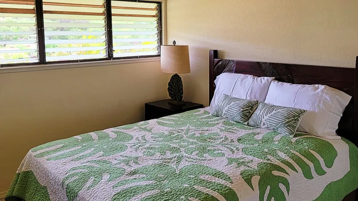 Bedroom with tropical decor: Green leaf-patterned quilt, white pillows, and two accent pillows. Wooden headboard and lamp....