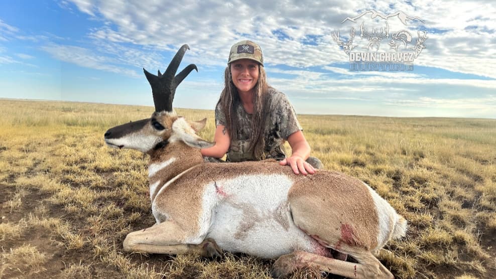 Woman in camo hat and shirt kneels next to a dead pronghorn in a grassy field. The sky is blue with clouds.