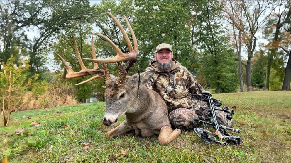 Man in camo kneels next to a large buck with impressive antlers. A bow rests beside them on green grass with trees in back...