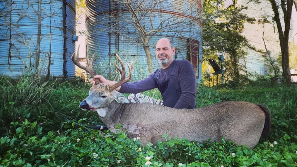 Man kneels next to a large deer with impressive antlers, both in tall grass. Two silos are visible in the background.